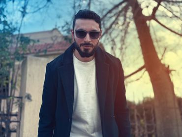 Man in sunglasses and coat posing outdoors near trees during sunset.