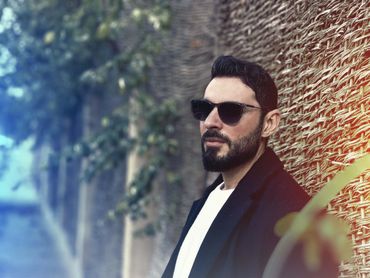 Bearded man wearing sunglasses leans against a textured wall outdoors.
