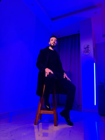 Man in black outfit sitting on a wooden stool in a blue-lit room.
