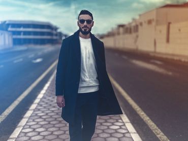 Stylish man walking confidently on a city sidewalk wearing sunglasses and a dark coat.