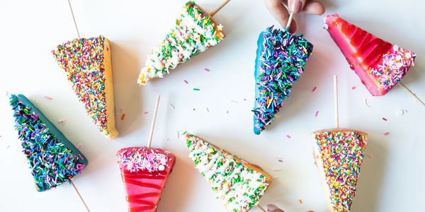 Colorful cake slices on sticks with sprinkles, held by two hands.