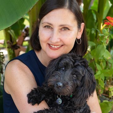 A smiling woman hugs her fluffy black dog outdoors among green plants.