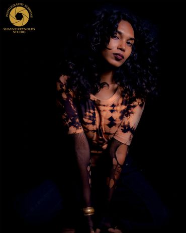 Woman with curly hair poses in dramatic lighting wearing a tie-dye top and fishnet sleeves.