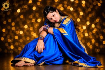 Woman in a vibrant blue sari sitting gracefully against a sparkling golden backdrop.