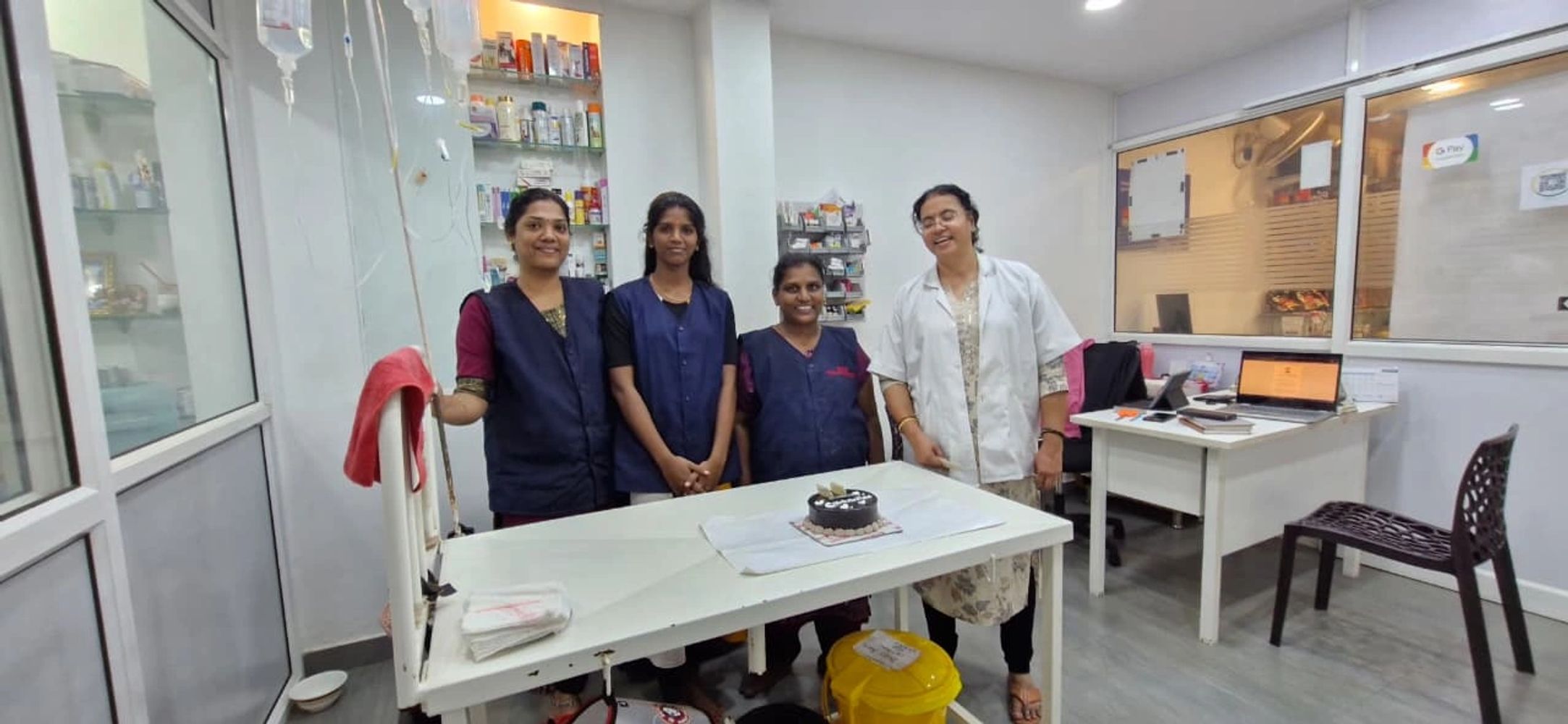 Four healthcare workers stand smiling around a table with a small chocolate cake in a clinic.