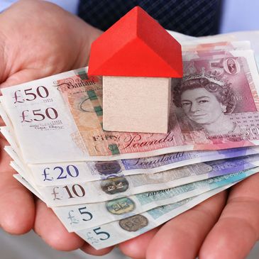 Hands holding British pounds with a small wooden house model.