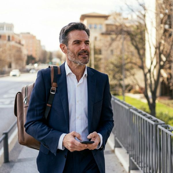 Man in suit walking outdoors with backpack and phone.