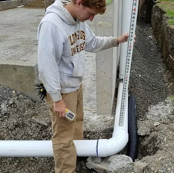 A worker measures a section of white PVC pipe in a trench with a vertical measuring pole.