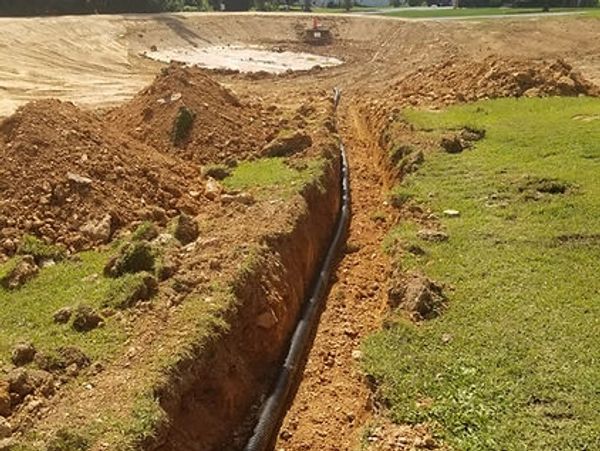 A narrow, deep trench with a black corrugated pipe running through it, leading to a large, empty pit