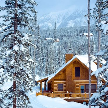 Cozy wooden cabin surrounded by snowy pine trees and mountains.