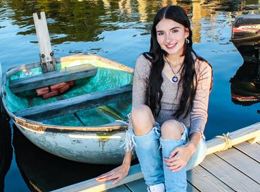 A young woman with long black hair sits on a dock beside a small boat.