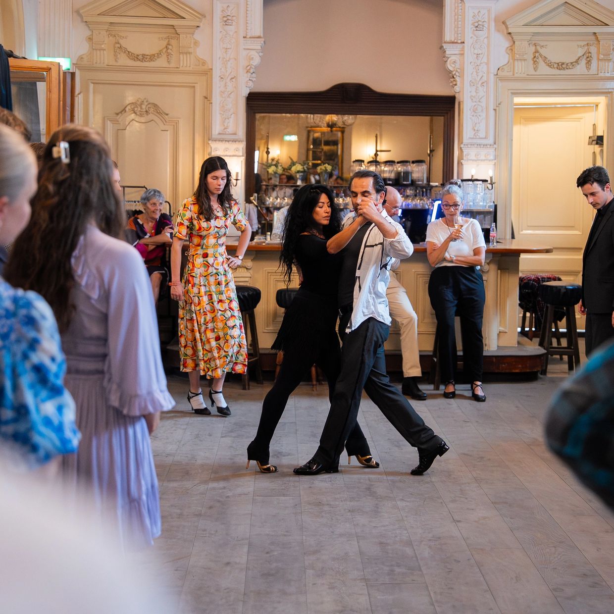 A couple dances tango in a vintage room surrounded by onlookers.