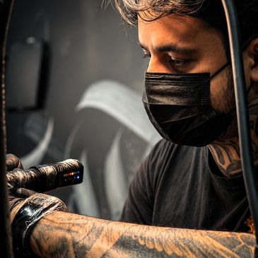 Tattoo artist wearing a black mask focused on tattooing.