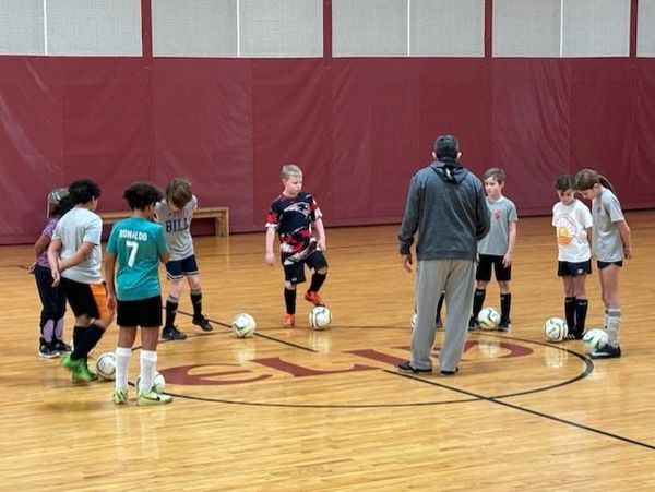 Children practicing soccer indoors under coach supervision.