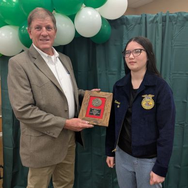 An older man presents an award plaque to a young woman in a blue jacket with FFA emblem.