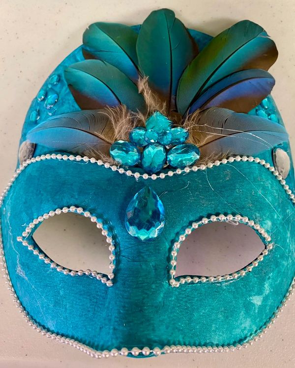 Turquoise masquerade mask adorned with feathers and gems.
