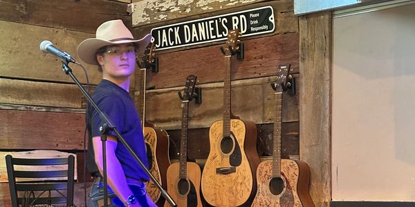 A cowboy-hat wearing man stands on a rustic stage with guitars and a tip jar.