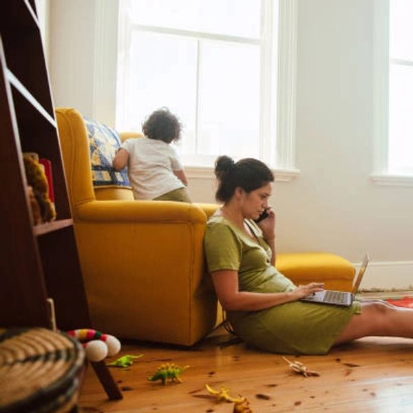 Woman working on laptop while child looks out window on yellow couch.