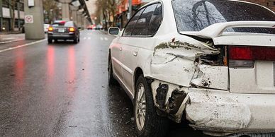 White car with rear-end damage parked on a wet city street.