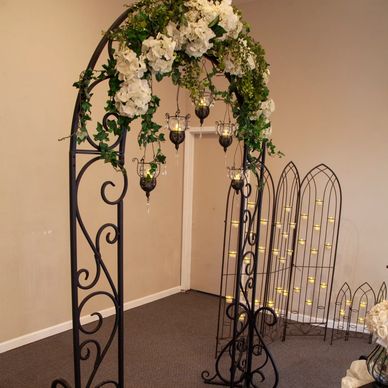 Elegant black metal arch decorated with white flowers and hanging candle holders.