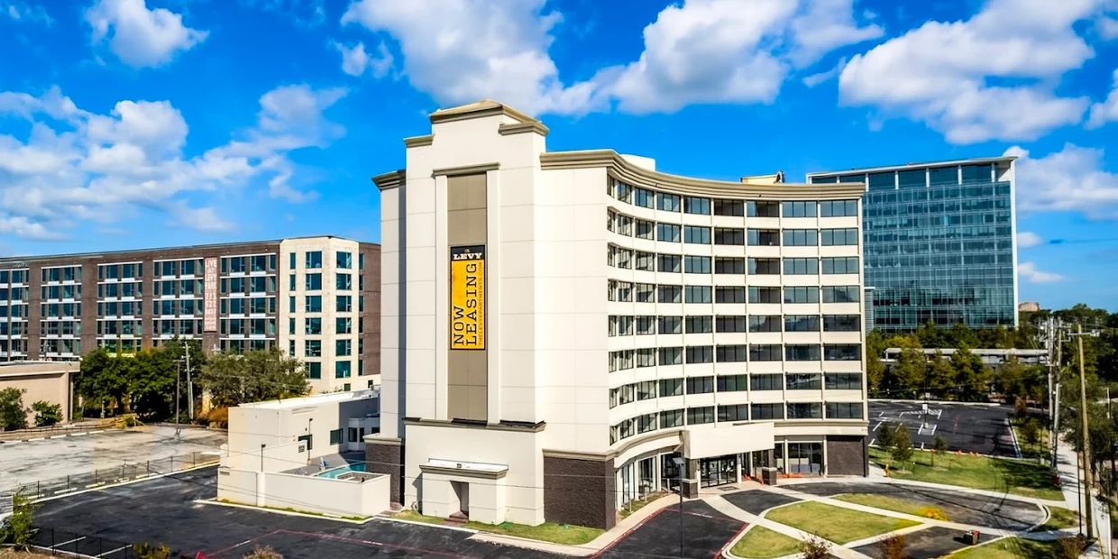 Modern apartment complex with a "Now Leasing" sign under a partly cloudy blue sky.