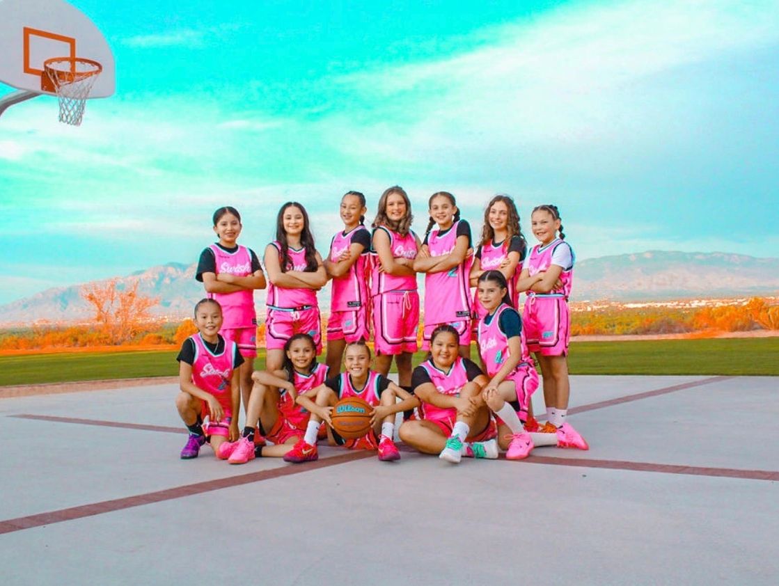 A girls' basketball team poses confidently on an outdoor court with mountains in the background.