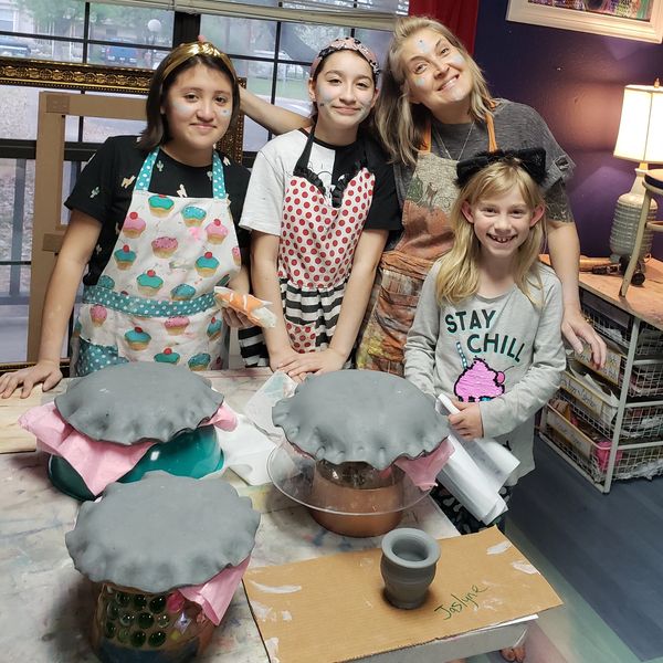 Four people smiling in a pottery class with clay-covered projects on the table.