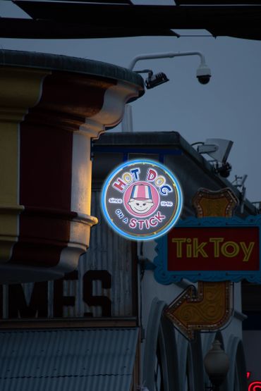 Neon sign for "Hot Dog on a Stick" glowing at dusk near a vintage-style building.