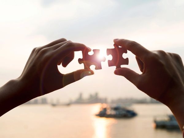Two hands holding puzzle pieces together against a sunset.
