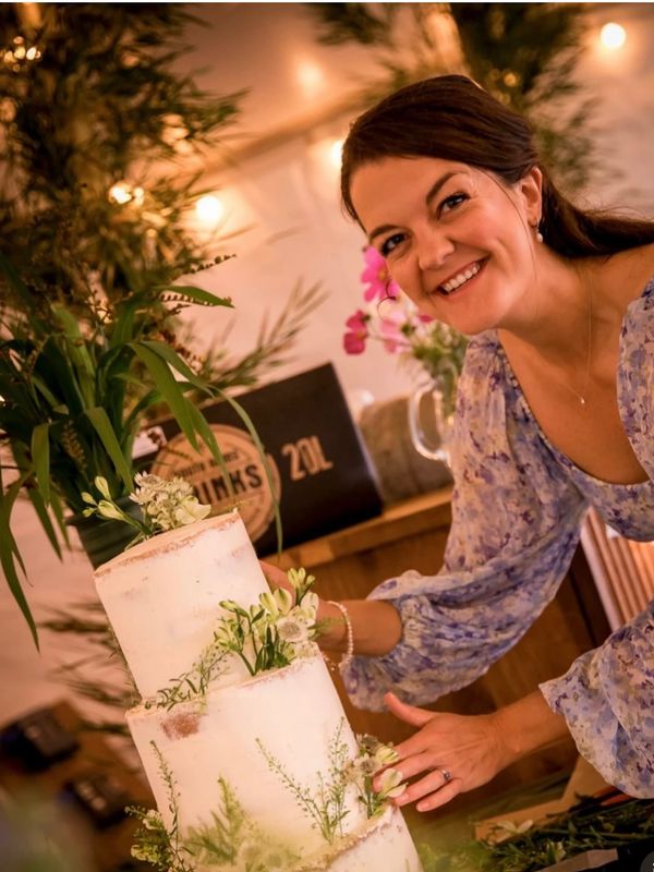 A smiling woman in a floral dress decorates a three-tier white cake with greenery.