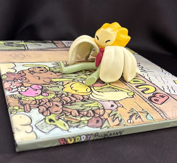 A whimsical banana character sits on a colorful illustrated book titled 'Buddy Business'.