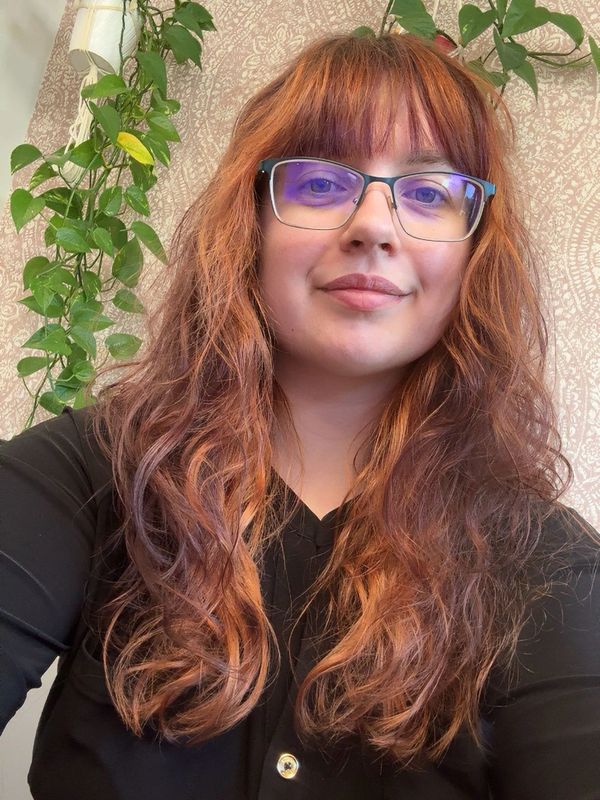 Young woman with wavy red hair and glasses smiling indoors.