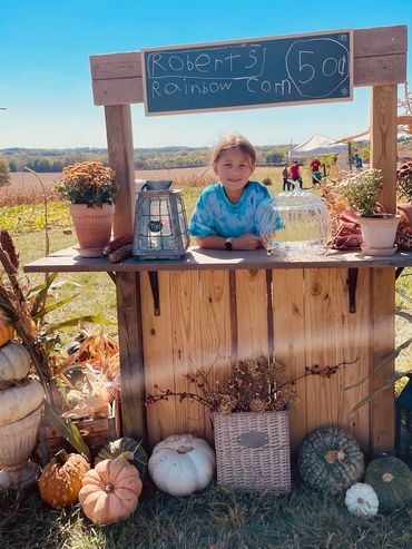 Young child selling rainbow corn for 50 cents at a rustic outdoor stand.