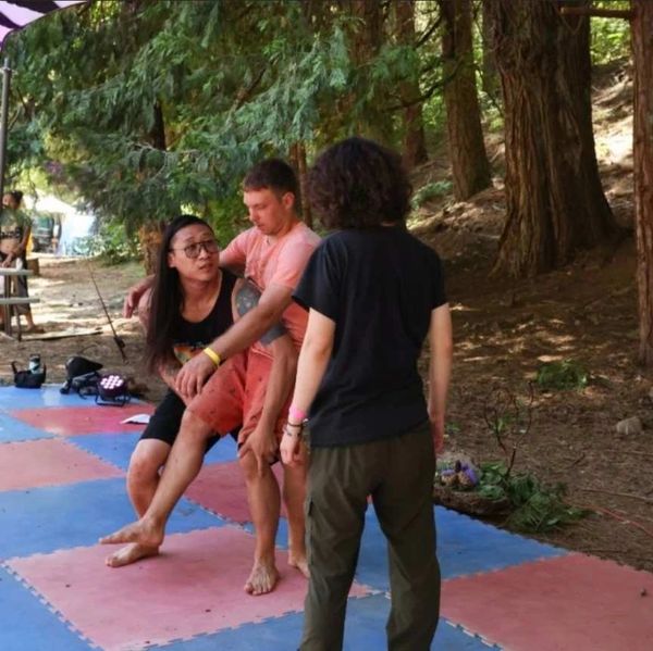 Three people practicing self-defense or martial arts outdoors on mats under trees.