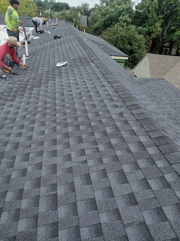 Workers installing new gray asphalt shingles on a large roof under a cloudy sky.