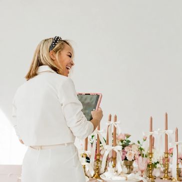 Woman in white holding a tablet, smiling by a decorated table.