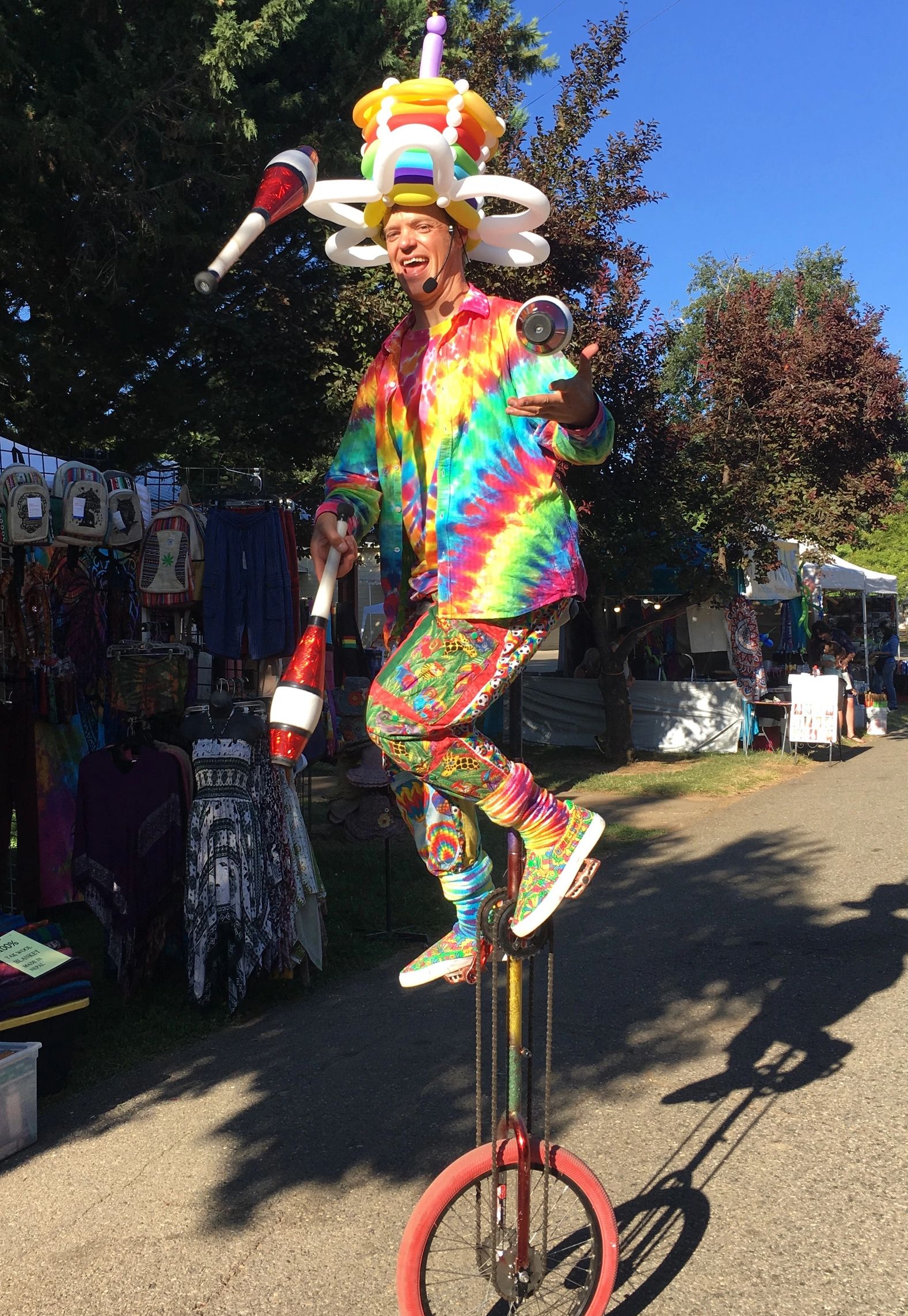 Jeremy the Juggler juggling clubs on giraffe unicycle at county fair.  Tie-dye outfit.