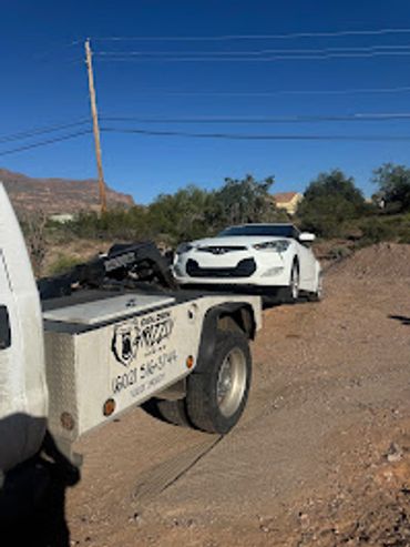 A damaged white car being towed on a flatbed truck in a rural area.