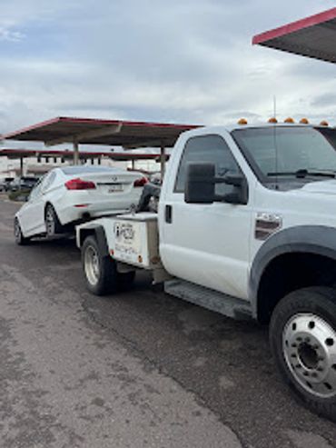 A white tow truck is transporting a white car in a parking lot with covered spaces.