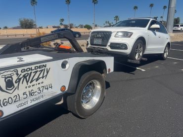 White Audi SUV being towed by Golden Grizzly Towing truck on a sunny day.