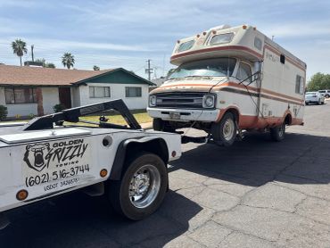 An old camper van being towed by a Golden Grizzly tow truck on a sunny day.