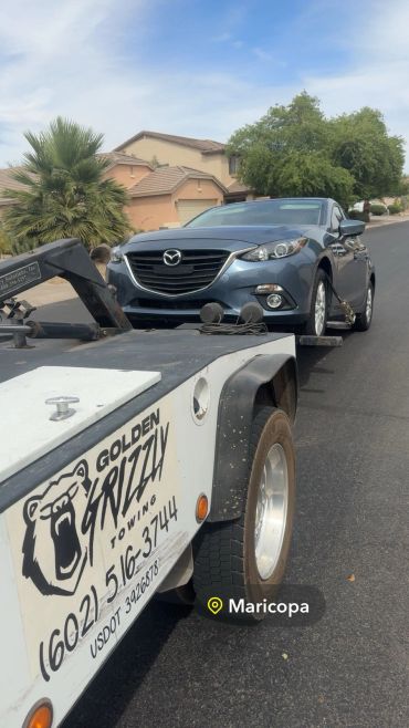 A blue Mazda car being towed by Golden Grizzly Towing in a residential area.