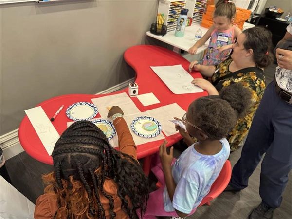 Children and adults engaged in a creative drawing activity at a red table.