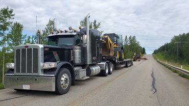 A black semi-truck hauling a large yellow excavator on a flatbed trailer down a highway