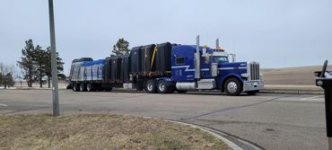 A clean blue, well-maintained Peterbilt semi-truck hauling miscellaneous goods on a stepdeck trailer