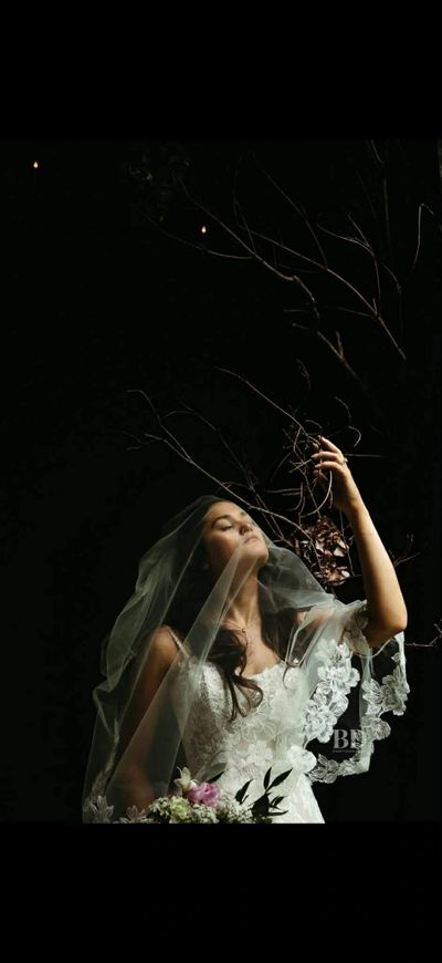 Bride in a lace wedding dress with veil, holding flowers, reaching toward bare branches.