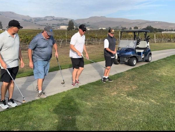 Four men preparing to play golf on a sunny course with a golf cart nearby.