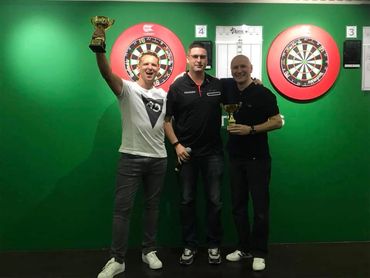 Three men celebrating with trophies in front of dartboards.