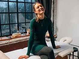 Smiling woman sitting on a massage table in a cozy room with large windows.