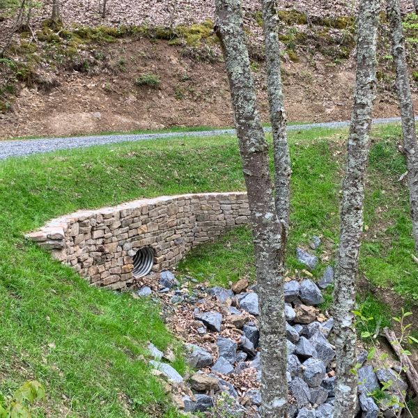 Stone culvert drains water under a gravel road in a green forest area.